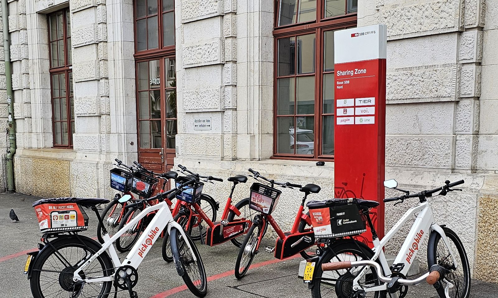 White Pick-e-Bike S-pedelecs and red PubliBike Velospot pedelecs parked next to a red Sharing Zone sign in front of the Basel SBB station building